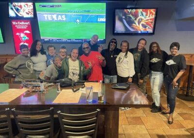 A diverse group of people stands and smiles around a long table in a sports bar, posing for a group photo with TVs showing a soccer match in the background.