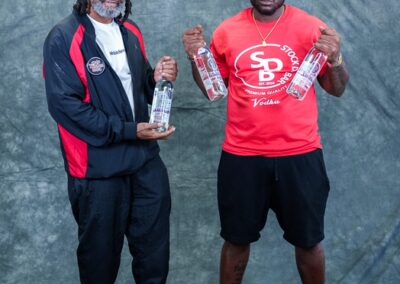 Two men stand side by side posing with bottles of vodka, one in a red shirt and cap, the other in a jacket and sunglasses against a textured backdrop.