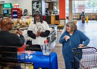 In a grocery store, a server pours samples from bottles on a blue-table setup while a shopper with a cart watches.