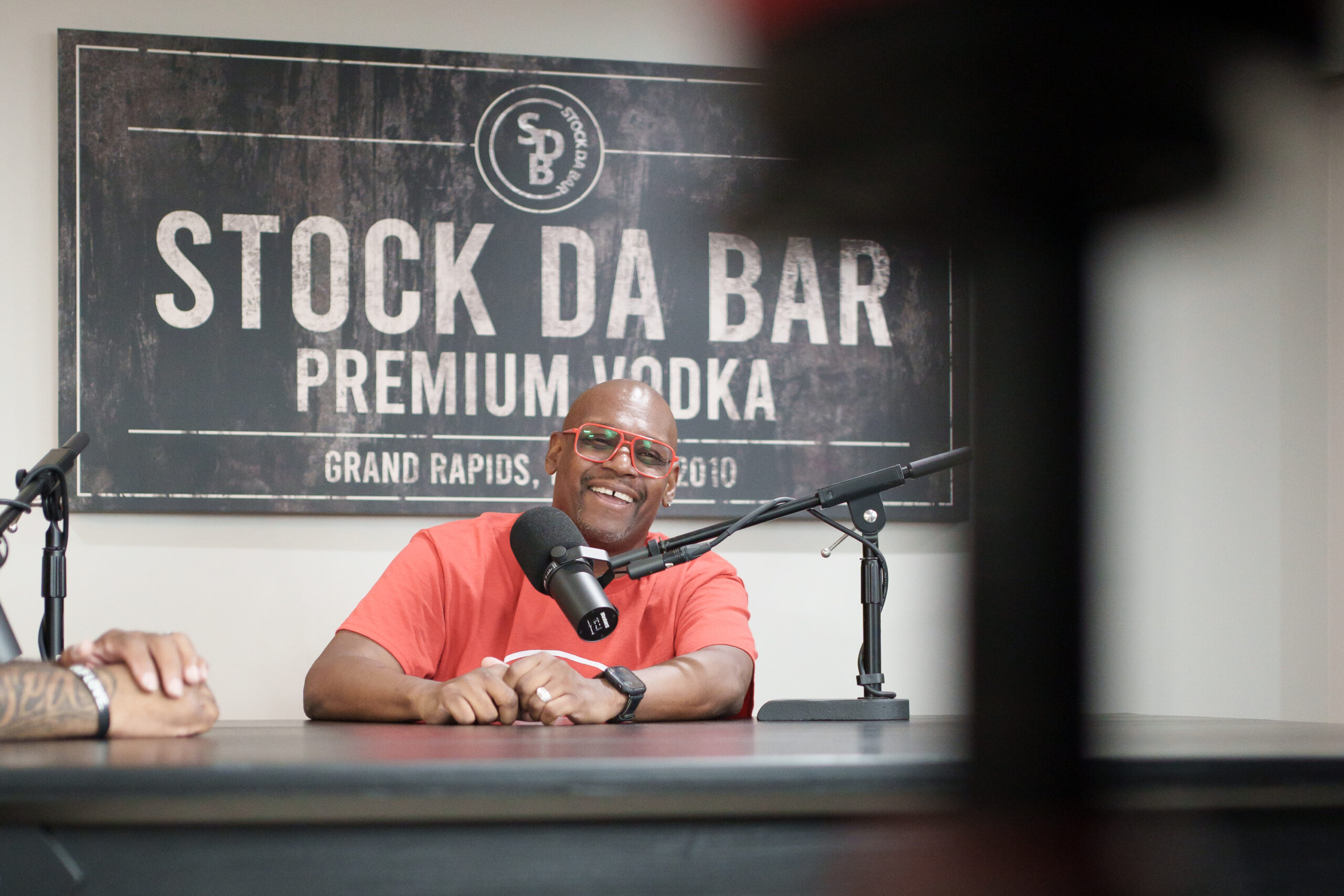 Smiling man in a red shirt and red glasses sits at a podcast mic setup with a large 'STOCK DA BAR' sign behind him.