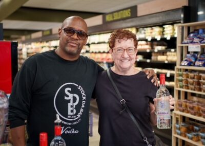 Two people stand together in a store near the liquor section; the woman holds a bottle while the man smiles with his arm around her shoulder, both looking at the camera.