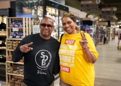 Two friends posing and smiling in a supermarket aisle, the man wearing a black T-shirt with a logo and the woman in a bright yellow shirt flashing a peace sign.