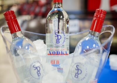 Three clear vodka bottles with red caps chilling in a transparent ice-filled bucket on a store counter, labels showing Vodka and blue logos.