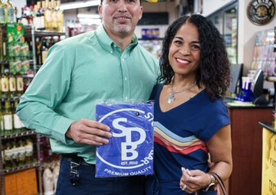 Smiling couple stands in a liquor store aisle; man in a green shirt and cap holds a blue SB sign, while woman in a blue striped dress holds a brown purse.