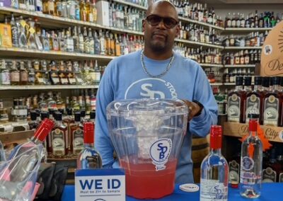 Man in a blue sweatshirt behind a sampling table with a blender and bottles in a liquor store.
