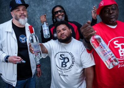 Four men posing with vodka bottles toward the camera; front‑center man wears a white T‑shirt with a circular logo and 'Vodka' text, others in casual hats and shirts against a dark backdrop.