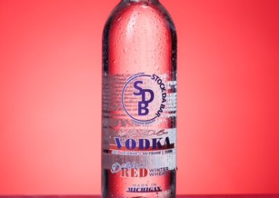 Clear glass vodka bottle with red cap and condensation, standing on a red surface with a red-to-pink gradient background; label features a blue SPB logo and the word VODKA.