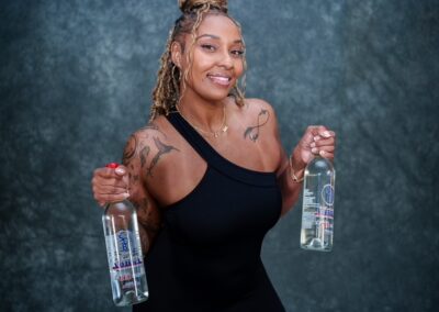 Smiling woman in a black one-shoulder top holds two clear bottles of liquor, tattoos visible on arms, against a dark backdrop.