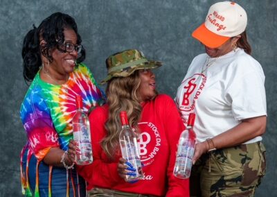 Three women smiling and toasting with clear bottles against a gray studio backdrop.