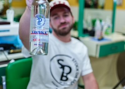 Man seated in a barbershop chair, holding a bottle of vodka toward the camera.