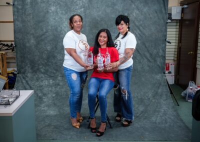 Three women in a photography studio pose together; the seated woman in red holds two bottles while the two standing women in white shirts touch her shoulders and smile.