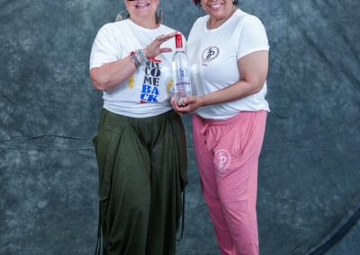 Two smiling women pose together against a textured blue backdrop; one in green pants and sunglasses, the other in pink joggers, holding a tall bottle between them.