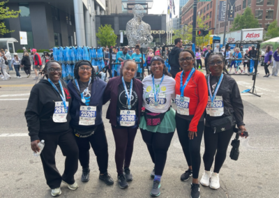 Seven women stand together on a city street, wearing race bibs and medals, posing after a race with a crowd and sculpture in the background.