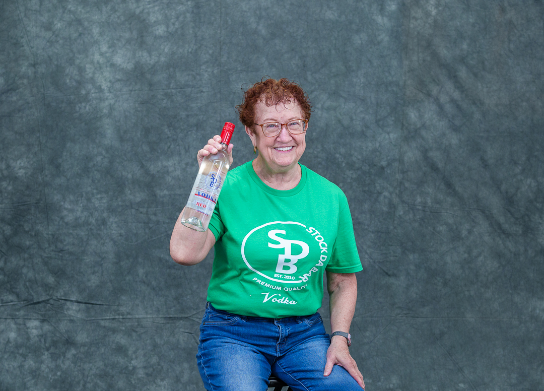 Smiling woman in a green shirt holds a bottle of vodka toward the camera against a gray backdrop.